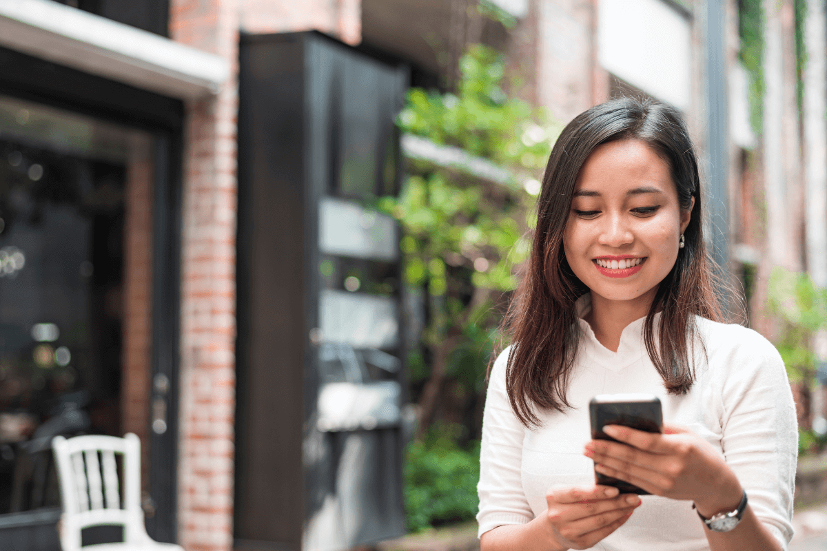 Usuário interagindo com busca local no celular Mulher sorrindo ao usar o celular na rua para buscar uma empresa local, ilustrando o comportamento do usuário e interação mobile.