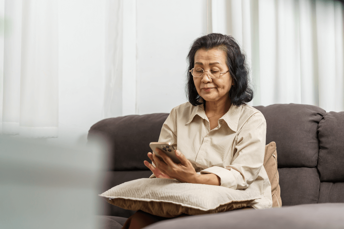 Mulher madura de óculos focada na tela do smartphone enquanto sentada no sofá, ilustrando a necessidade de acessibilidade e legibilidade em telas pequenas.