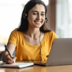 Mulher profissional sorrindo em home office com headset e notebook, ilustrando a busca por qual o primeiro passo para trabalhar na internet e iniciar uma carreira no mercado digital.
